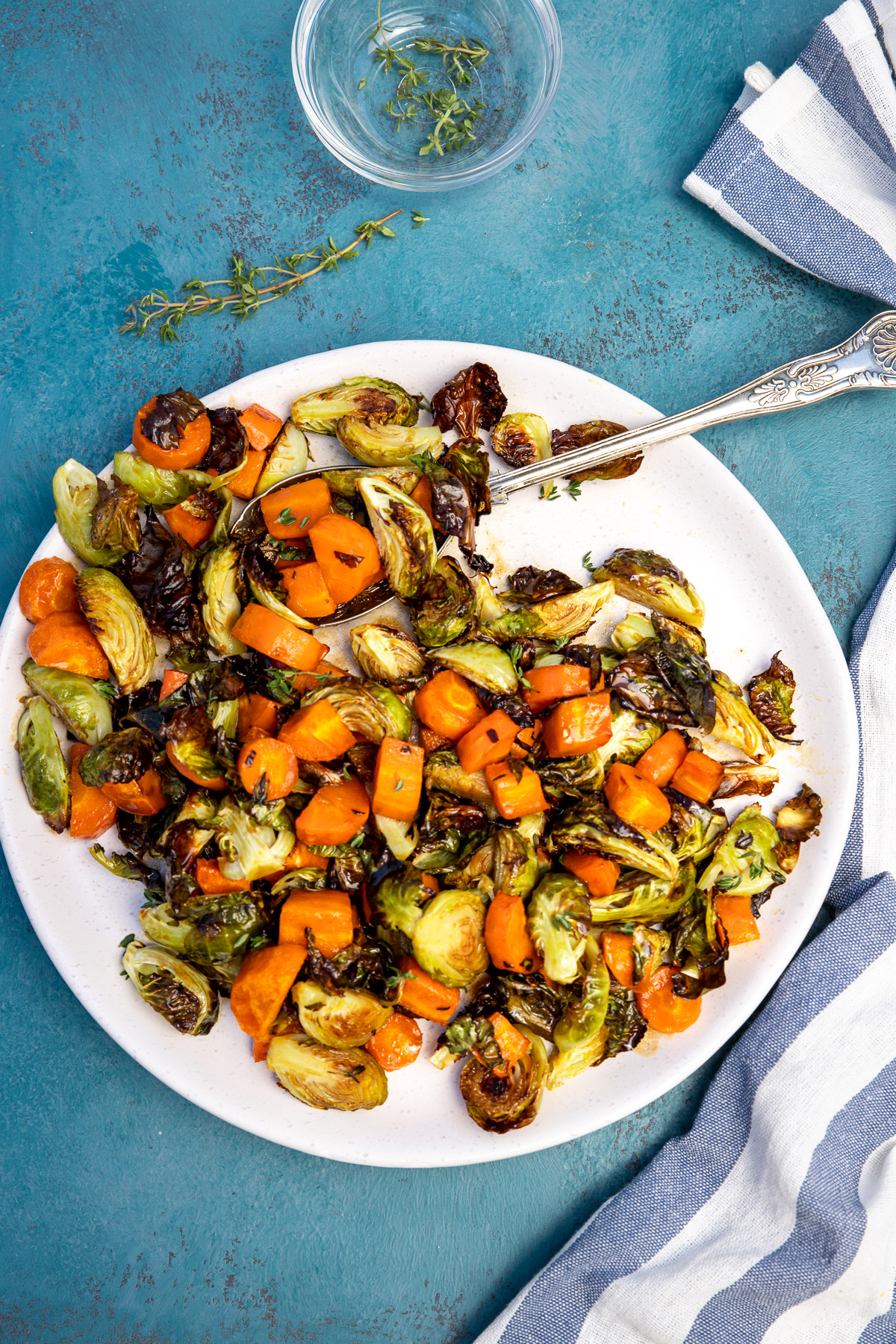 A plate of brussels sprouts and carrots with a spoon in it on a blue background with a blue striped tea towel