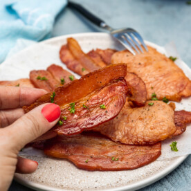 Someone holding up a piece of air fried bacon over a plate of it on a blue background with tea towel and a dish in the background