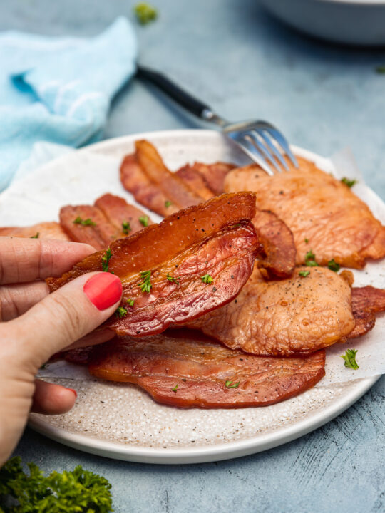Someone holding up a piece of air fried bacon over a plate of it on a blue background with tea towel and a dish in the background