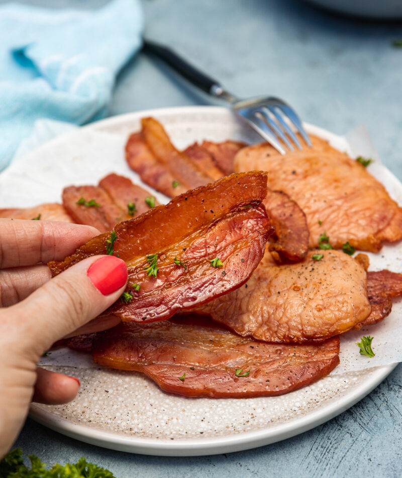 Someone holding up a piece of air fried bacon over a plate of it on a blue background with tea towel and a dish in the background