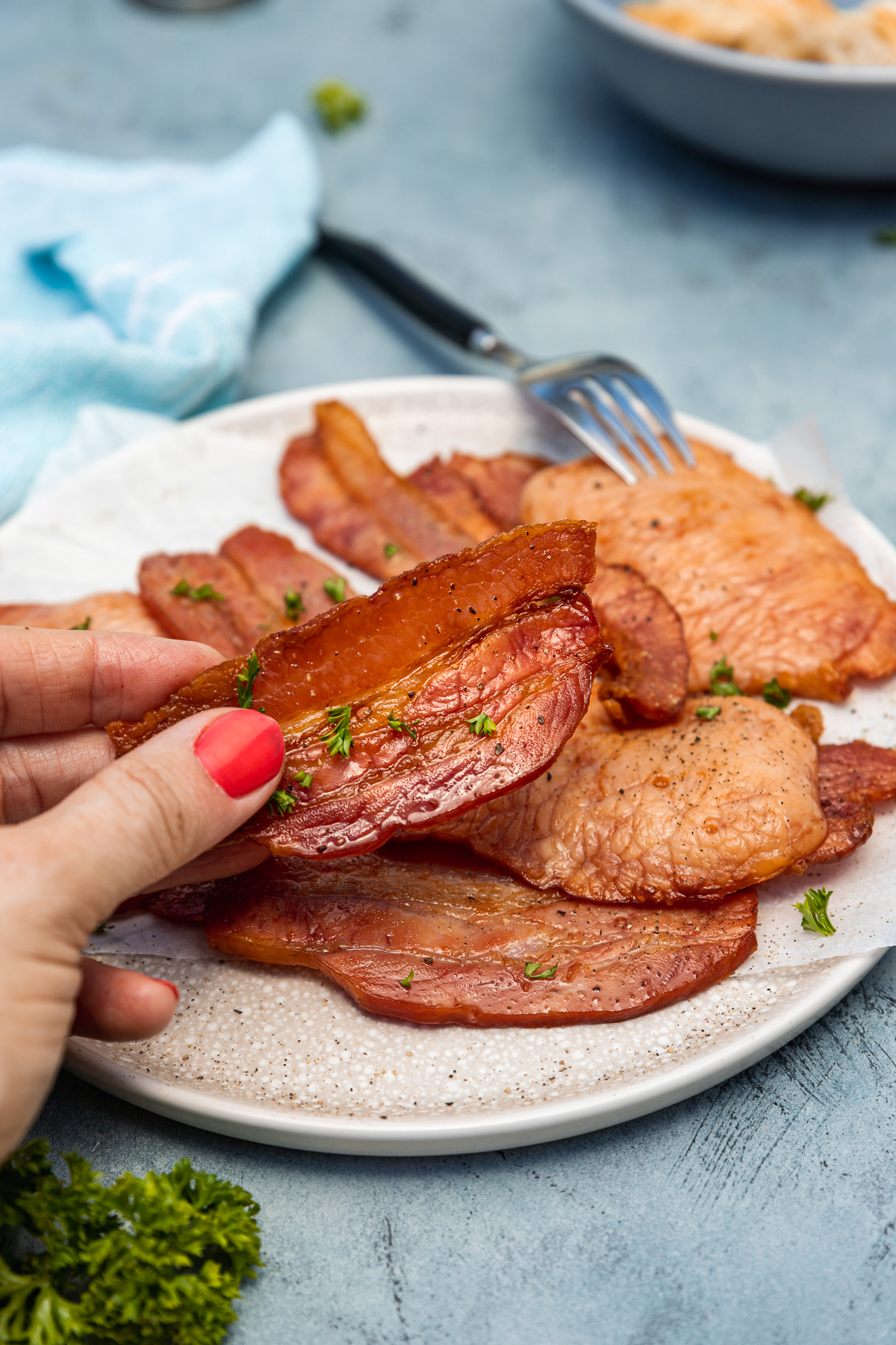 Someone holding up a piece of air fried bacon over a plate of it on a blue background with tea towel and a dish in the background