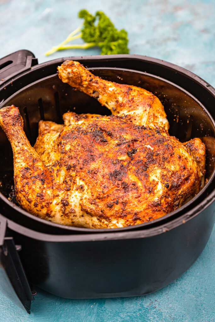 A whole cooked rotisserie style chicken in an air fryer basket on a blue background