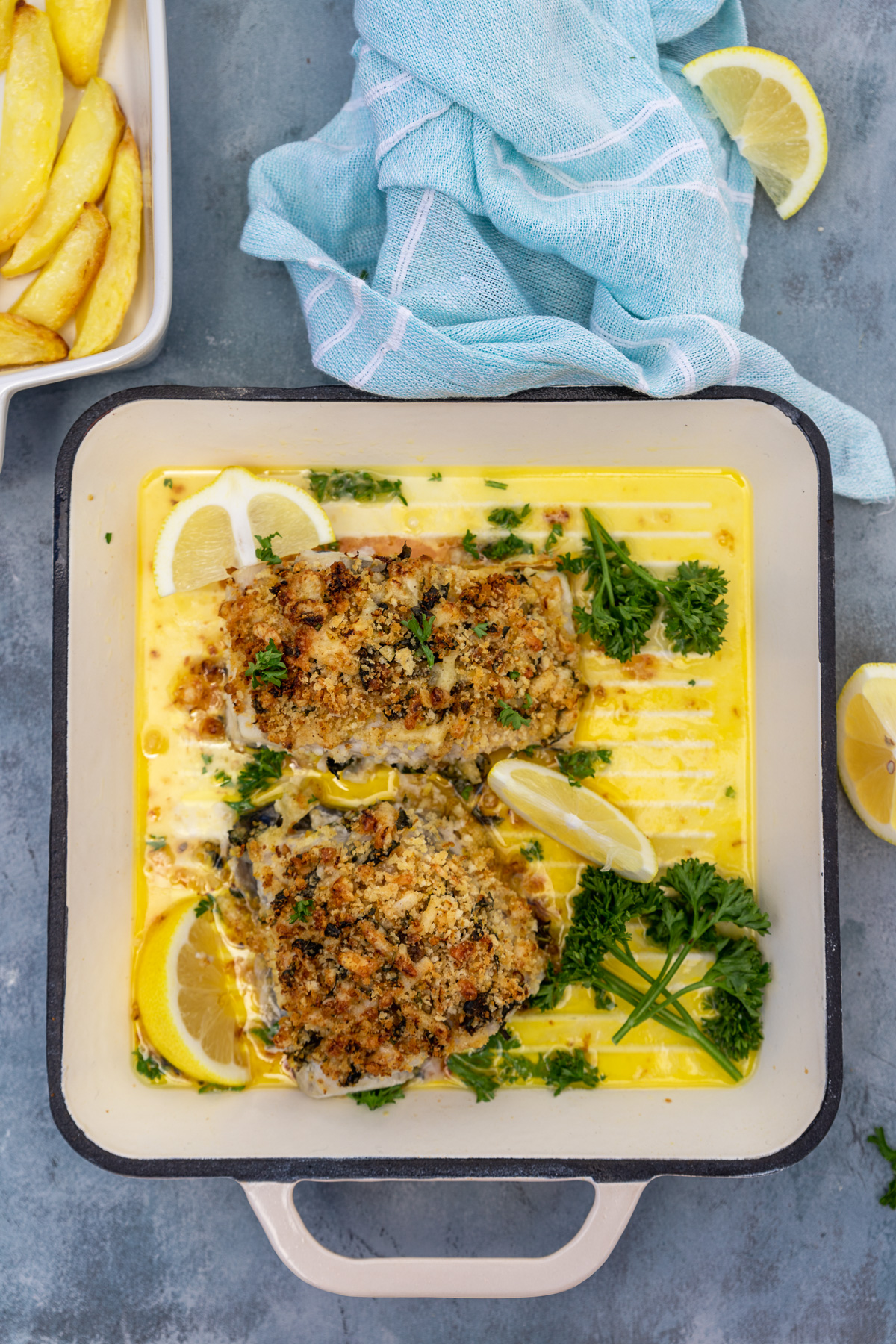baked cod with panko in a square shaped beige cast iron pan on a blue background  with blue tea towel from above