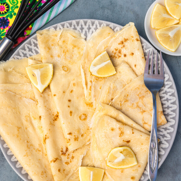 Overhead view of thin English pancakes on a grey and white plate and blue background with colourful tea towel, whisk, sugar and lemon