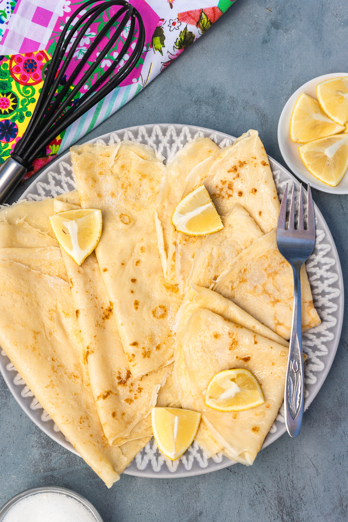 Overhead view of thin English pancakes on a grey and white plate and blue background with colourful tea towel, whisk, sugar and lemon