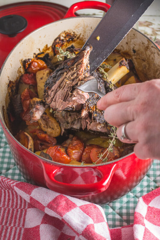 Closeup of someone's hands carving slow cooked Greek lamb and vegetables in a red cast iron pot.