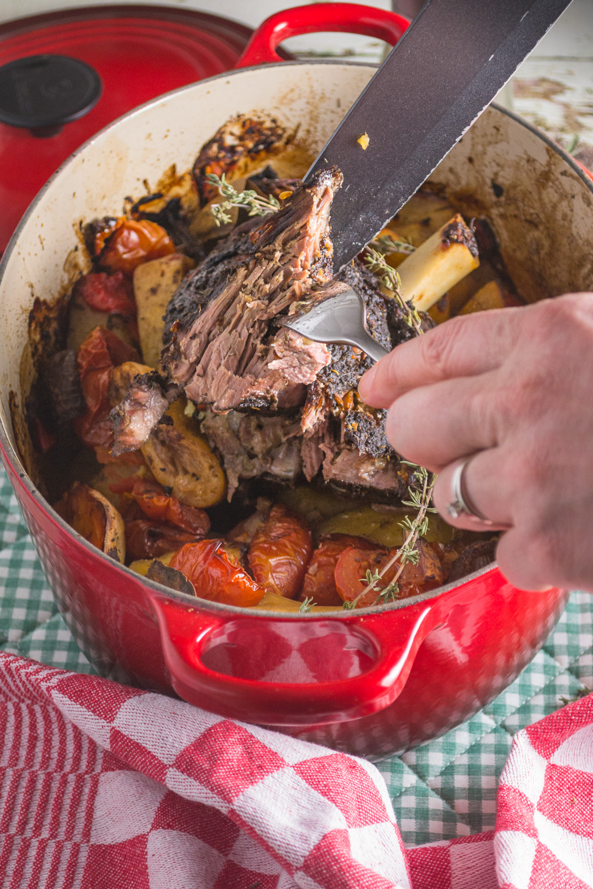 Closeup of someone's hands carving slow cooked Greek lamb on top of vegetables in a red pot