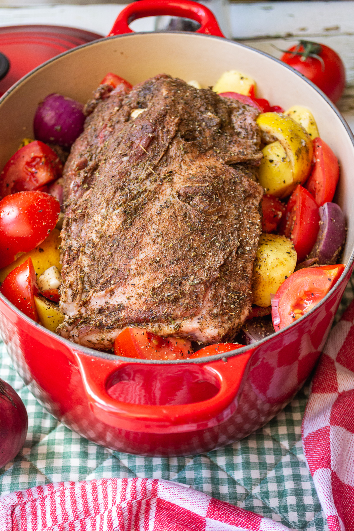 Raw lamb covered with a Greek rub on top of vegetables ready to be cooked in a red cast iron pot