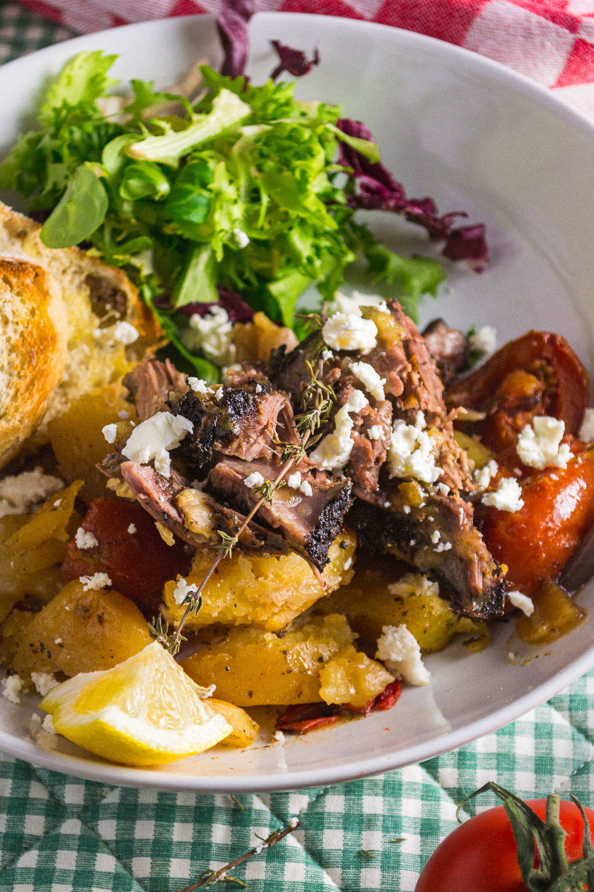 Closeup of Greek lamb kleftiko in a white bowl with garnishes including feta and green salad and bread