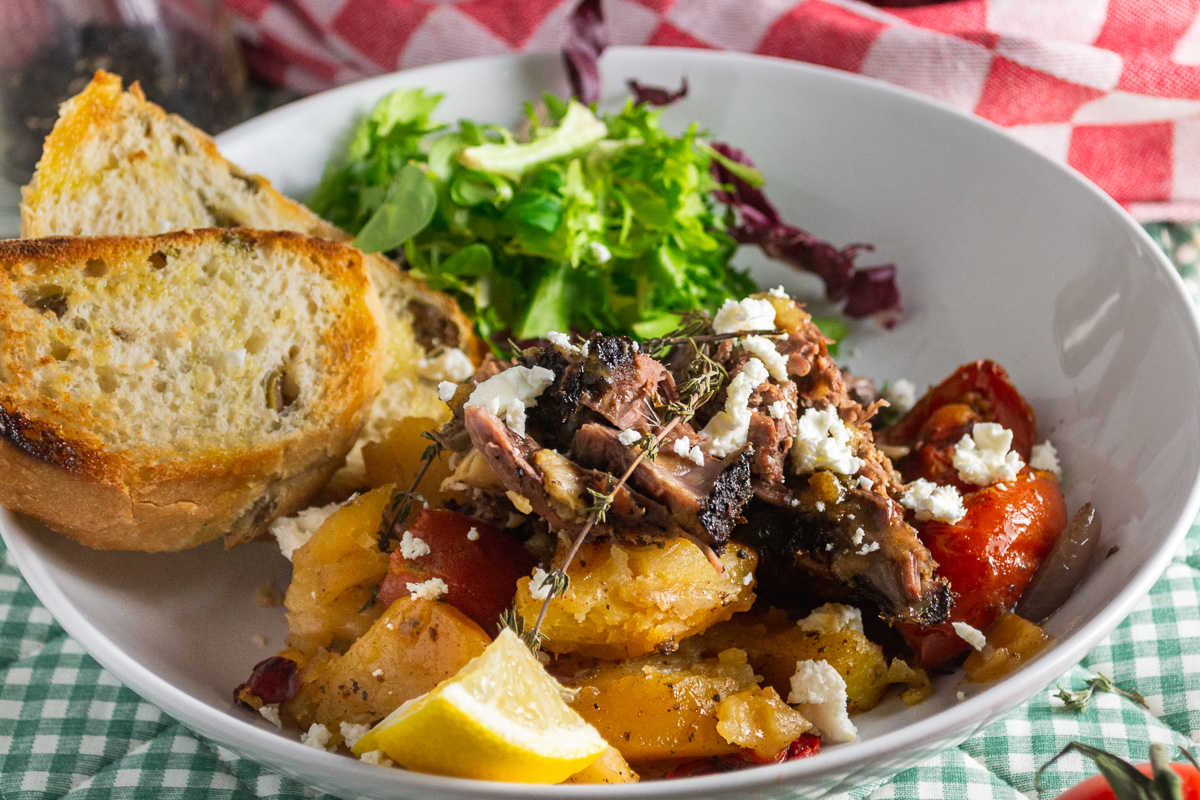 a closeup of a dish of Greek lamb and potatoes and vegetables with garnishes in a white dish and on a green and white checked background