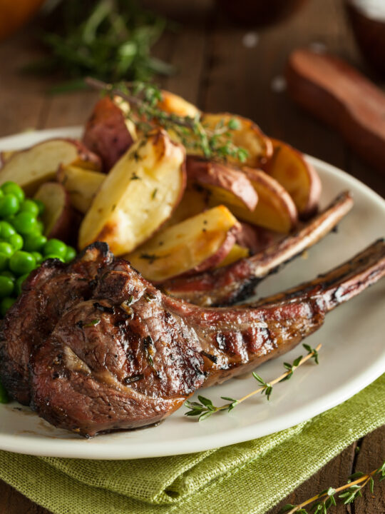 Closeup of grilled lamb chops with crispy potatoes and peas and thyme sprigs on a white plate and pale green napkin.