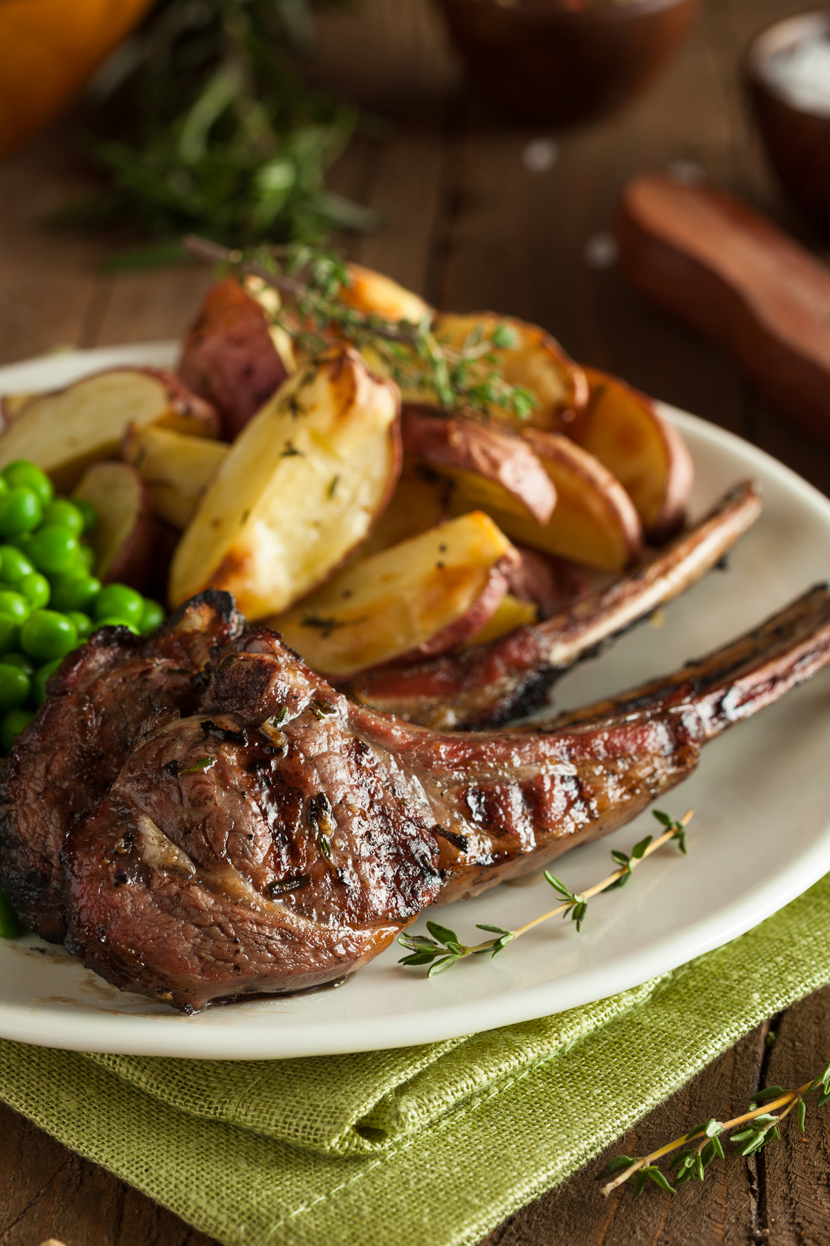 Closeup of grilled lamb chops with crispy potatoes and peas and thyme sprigs on a white plate and pale green napkin.