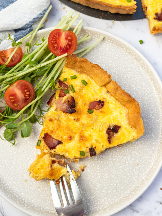 Slice of quiche and fork on a light beige plate with side salad photographed from above.