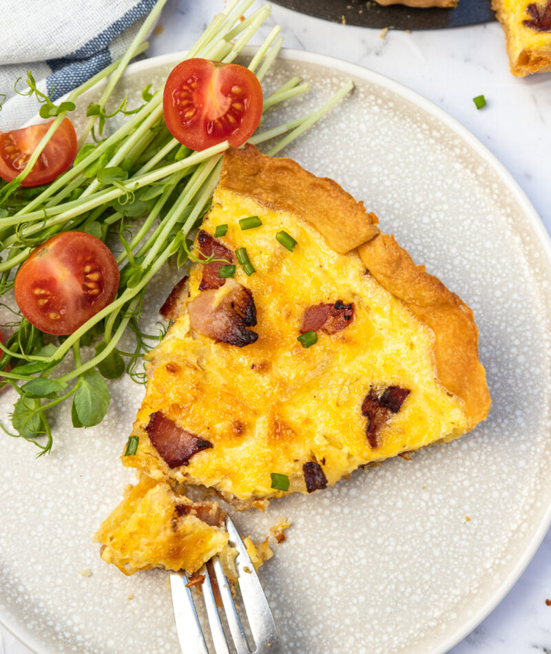 Slice of quiche and fork on a light beige plate with side salad photographed from above.