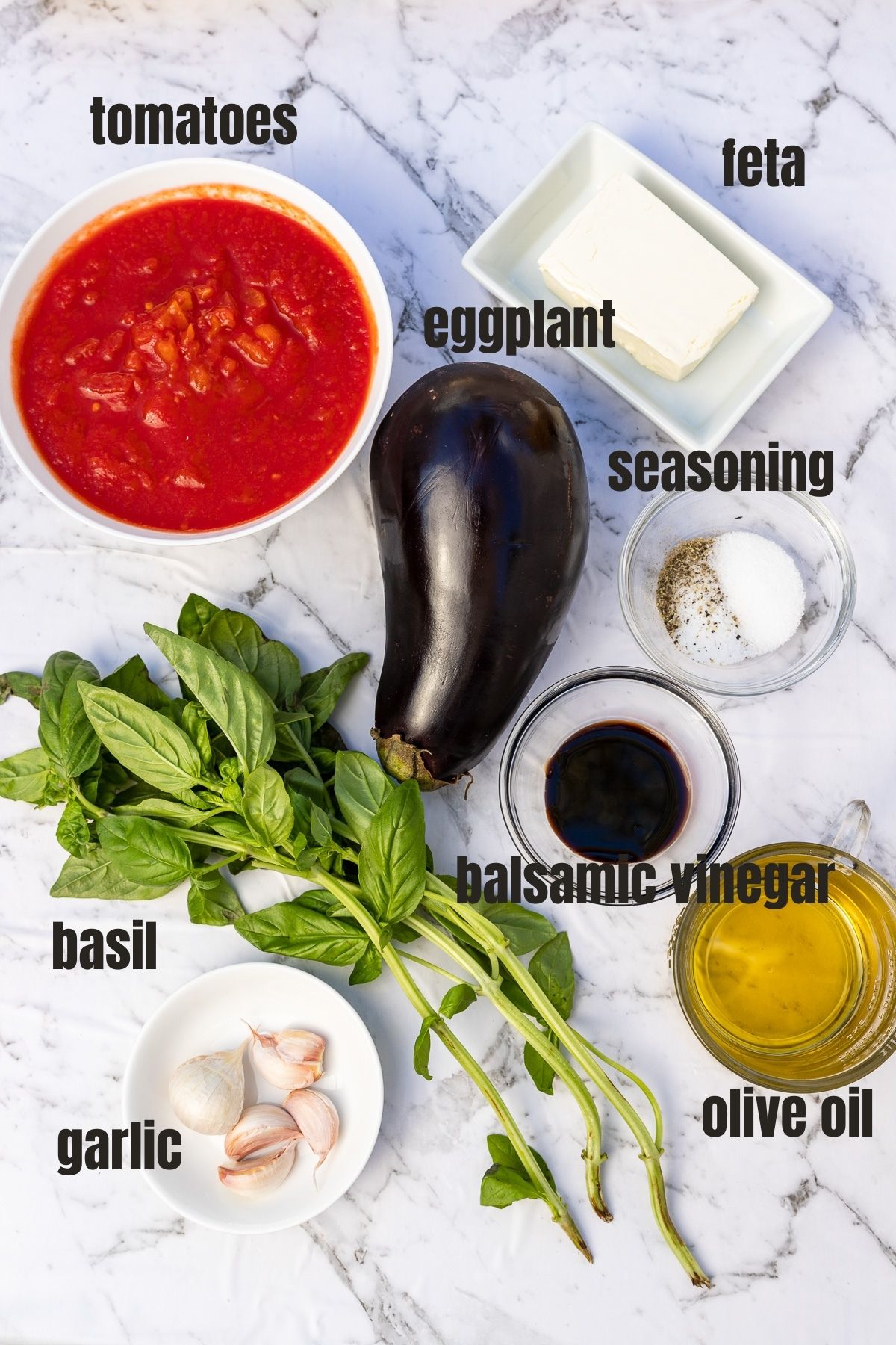 An overhead view of the ingredients for Greek baked eggplant, including eggplant, garlic cloves, olive oil, canned tomatoes, fresh basil, seasonings, feta and balsamic vinegar