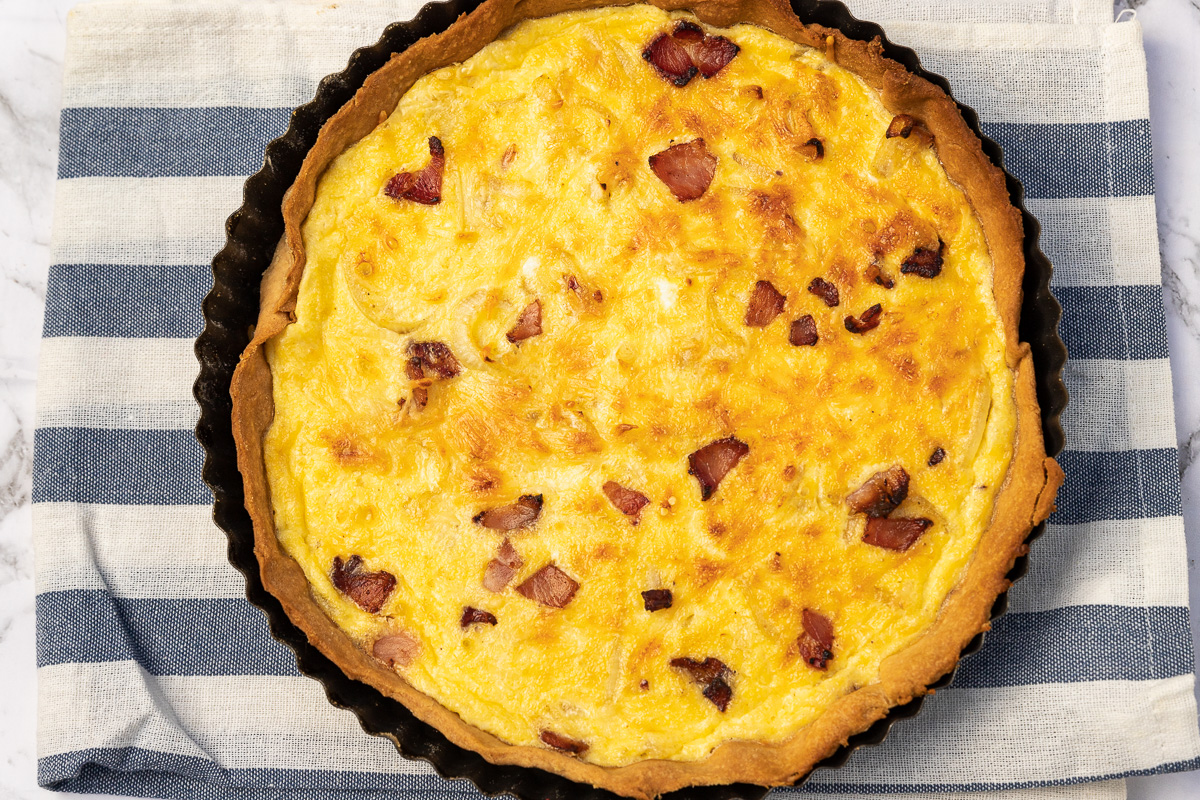 Whole cooked quiche lorraine from above on a white and blue striped tea towel and marble background.