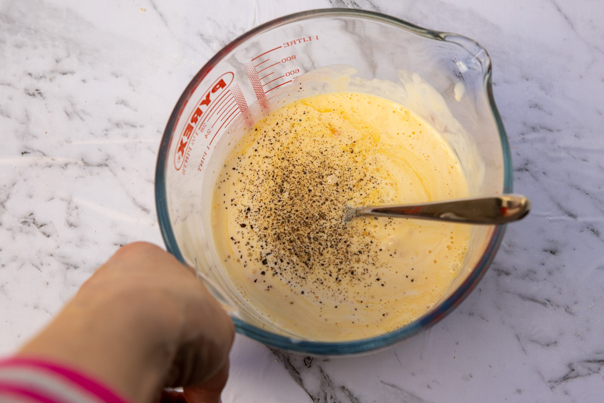 Someone holding a jug of egg custard mixture for quiche with a fork in it and on a marble background.