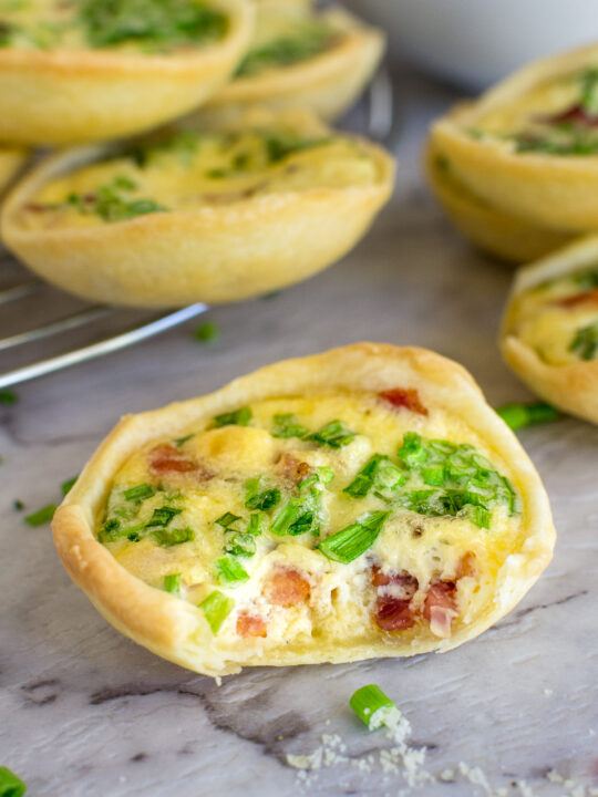 A closeup of a mini quiche lorraine with a small bite taken out of it on a marble background with more quiche in the background.