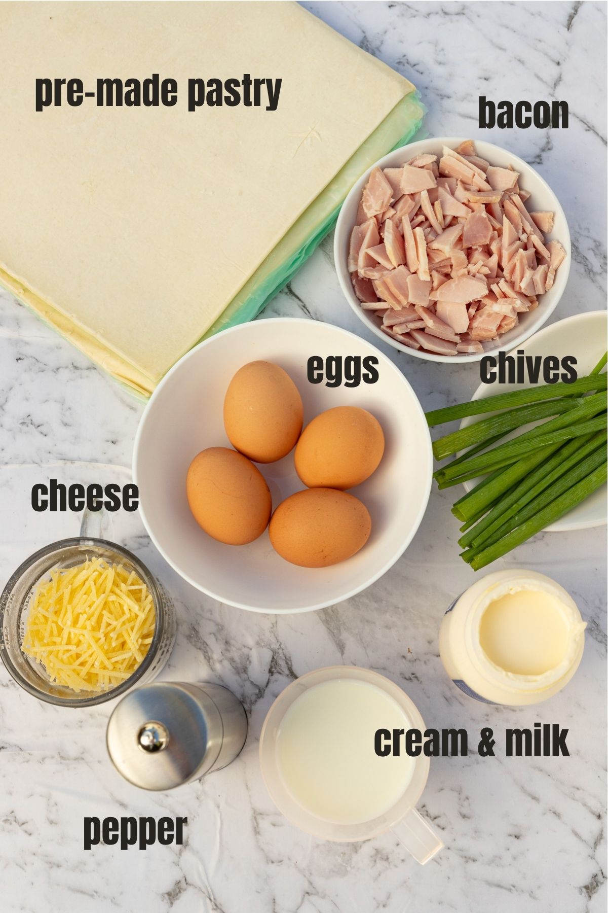 All the ingredients for mini quiche from above on a marble background including cream and milk, pepper, grated cheese, 4 eggs, chives, bacon and pre-made pastry.