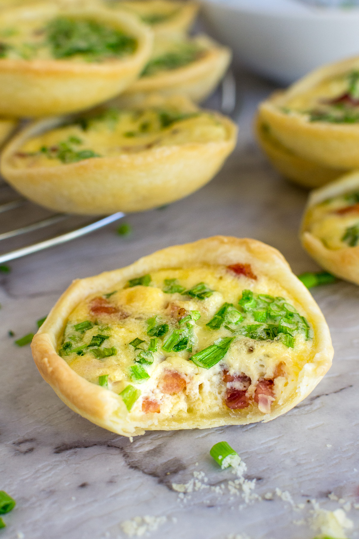A closeup of a mini quiche lorraine with a small bite taken out of it on a marble background with more quiche in the background.