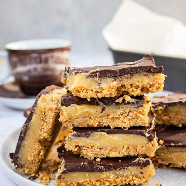 No bake caramel slice piled up on a light beige plate on a marble background with a teacup and baking pan in the background