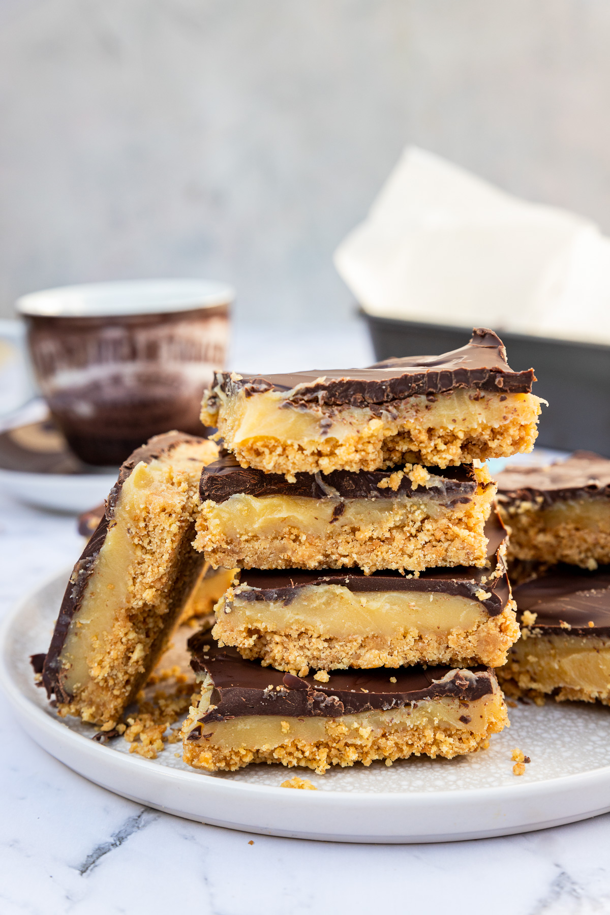 No bake caramel slice piled up on a light beige plate on a marble background with a teacup and baking pan in the background