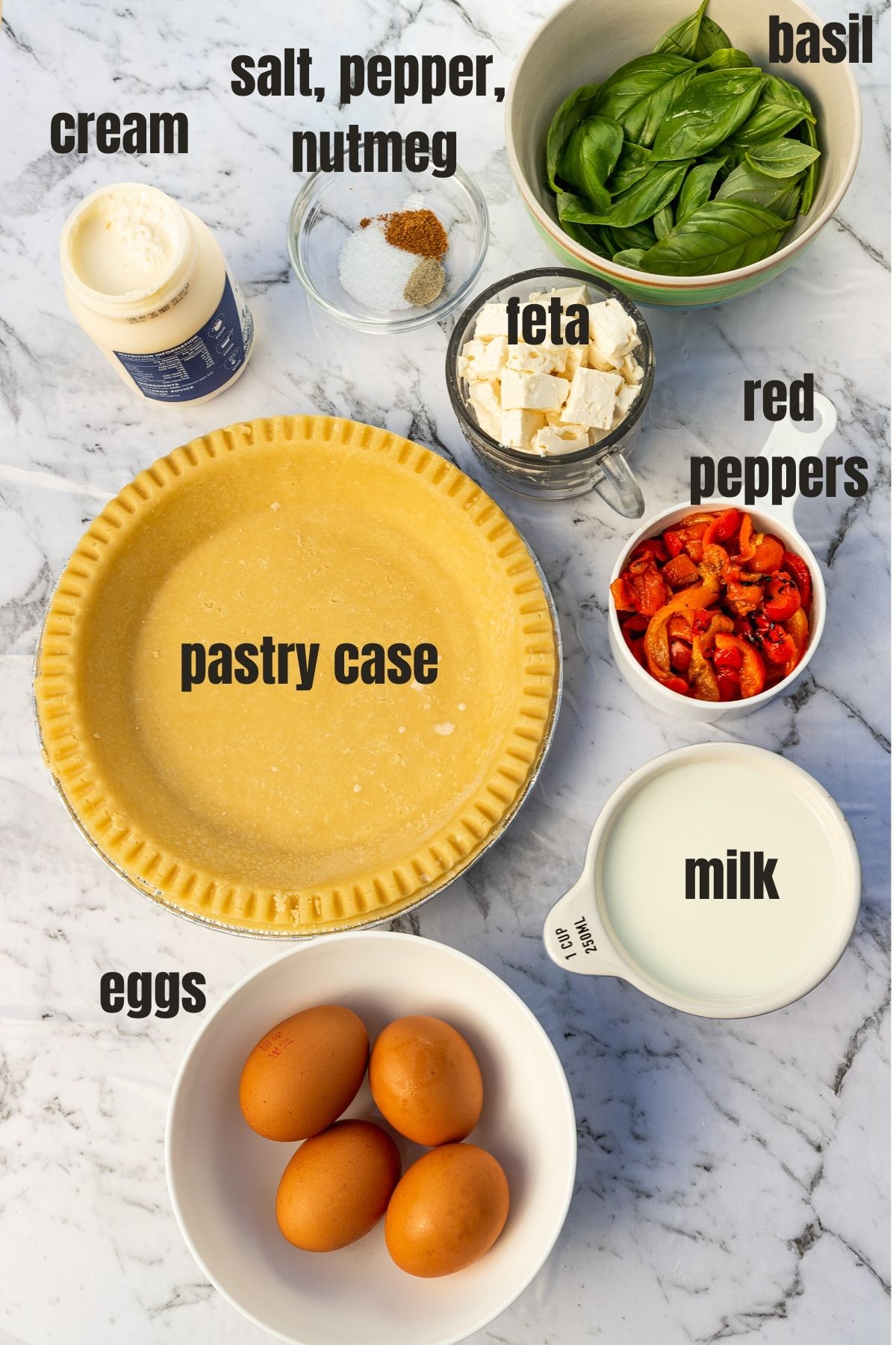 Overhead view of the labelled ingredients to make a red pepper feta quiche including eggs, milk, cream, basil, a pastry case, red pepper strips, feta cubes and seasoning.