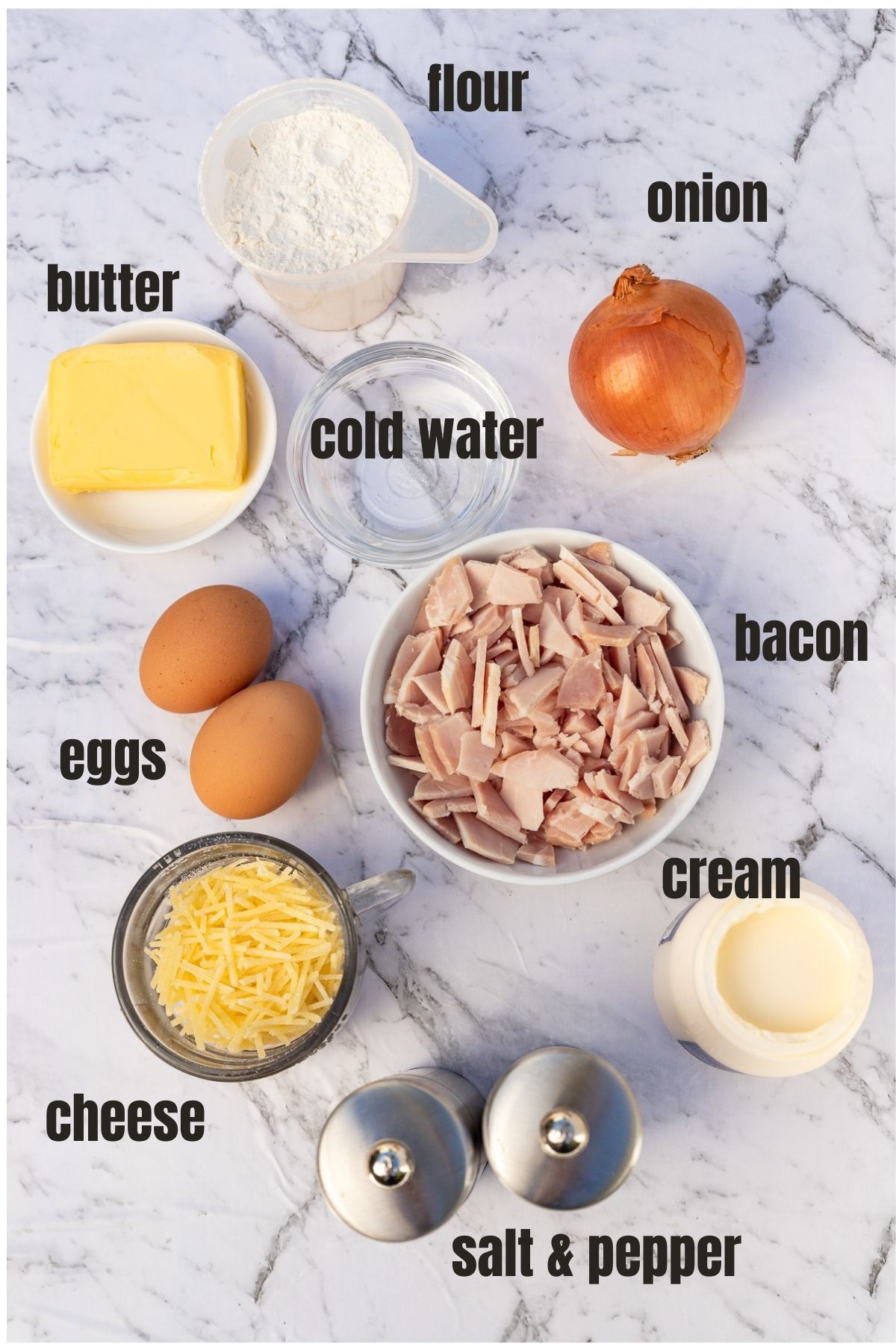 Overhead of all the ingredients to make a quiche lorraine including flour, butter, water, onion, eggs, cheese, seasoning, cream and bacon.