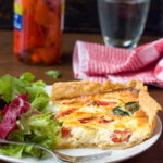 A square slice of homemade red pepper and feta quiche on a white plate with green salad and with ingredients and water and a red tea towel in the background.