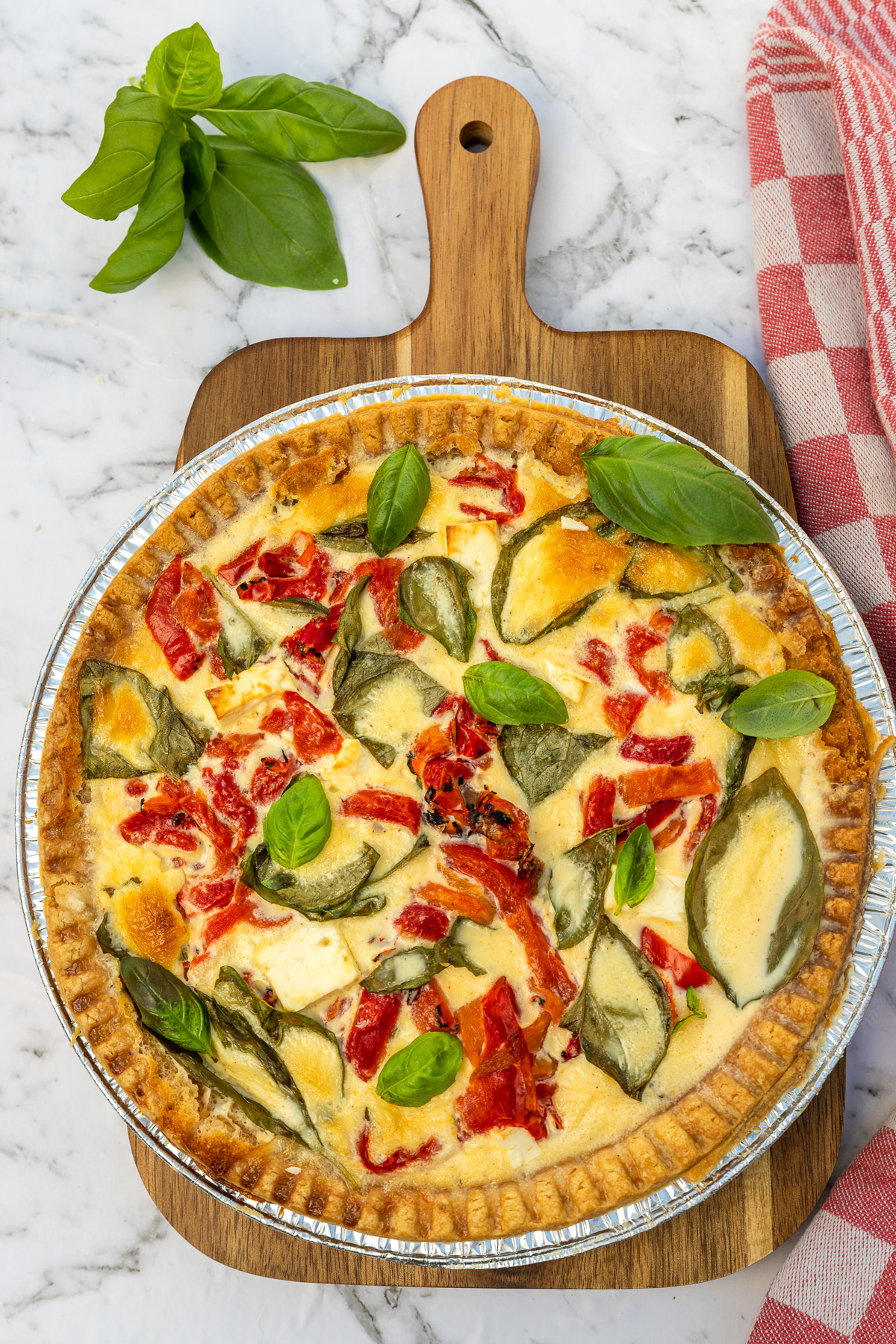 Overhead view of a colorful red pepper and feta quiche on a wooden board and marble background with a red checked tea towel.