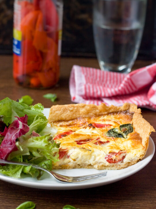 A square slice of homemade red pepper and feta quiche on a white plate with green salad and with ingredients and water and a red tea towel in the background.