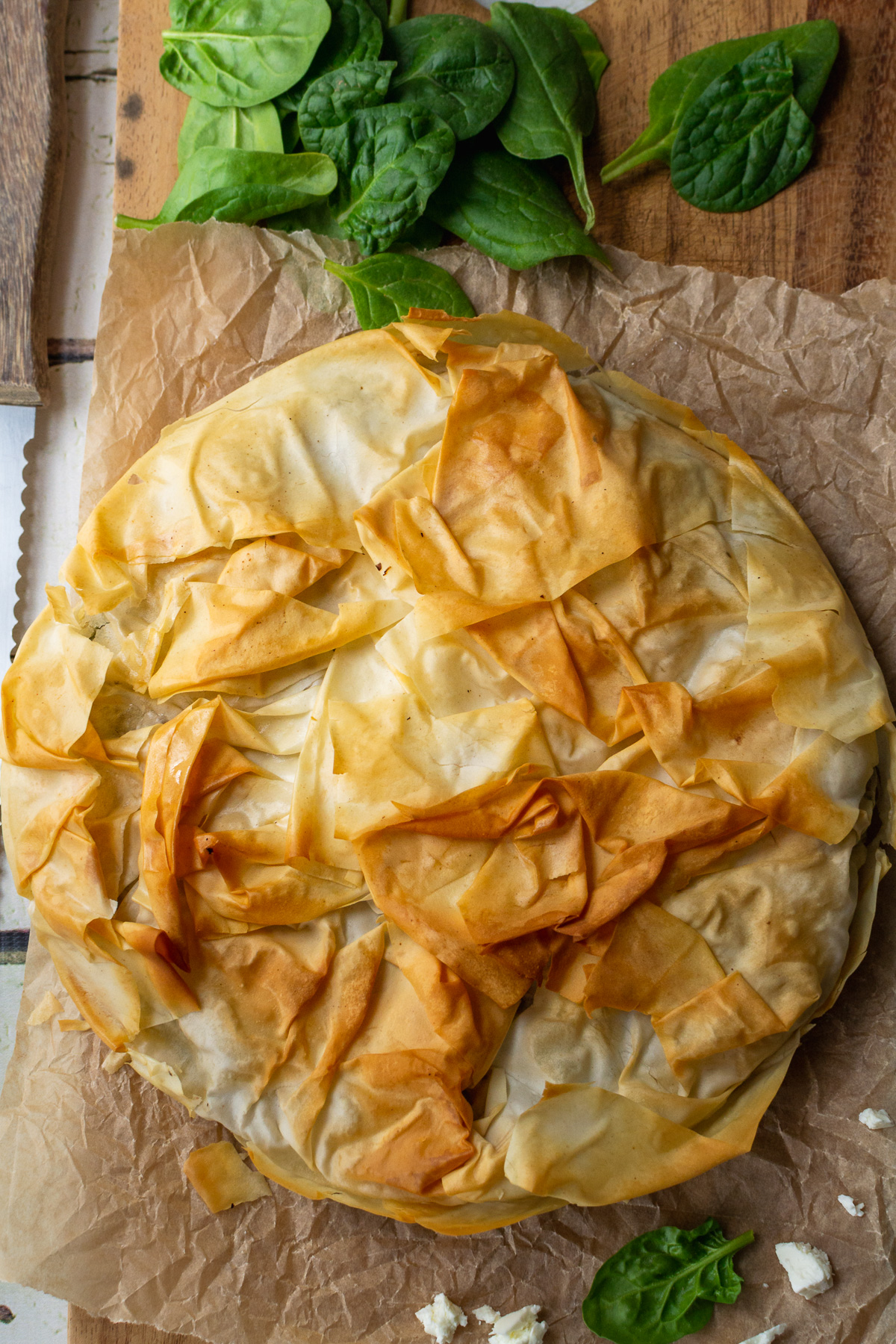 Overhead view of a Greek spinach and feta filo pie with a golden, crisp top on baking paper and a wooden board.