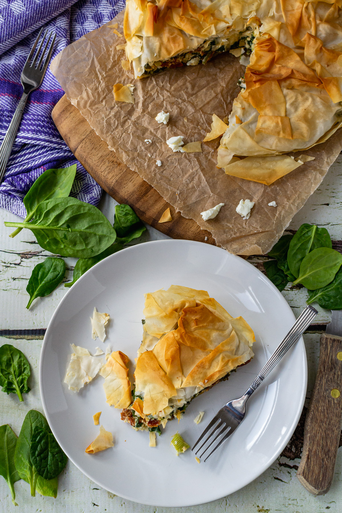 Overhead view of a slice of spinach feta pie on a white plate with a fork and with the rest of the pie in the far right corner on a wooden board and white wood table.