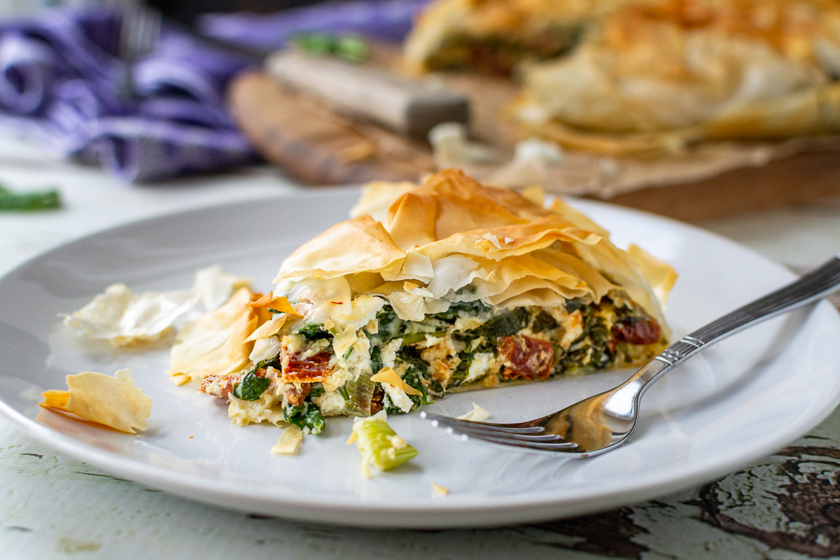 A closeup of a slice of spinach feta filo pie on a white plate with a fork.