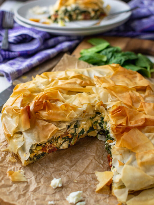 A round spinach and feta filo pie with a big piece cut out of it on baking paper and with a piece of the pie in the background.