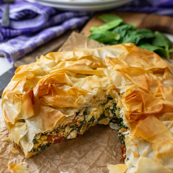 A round spinach and feta filo pie with a big piece cut out of it on baking paper and with a piece of the pie in the background.