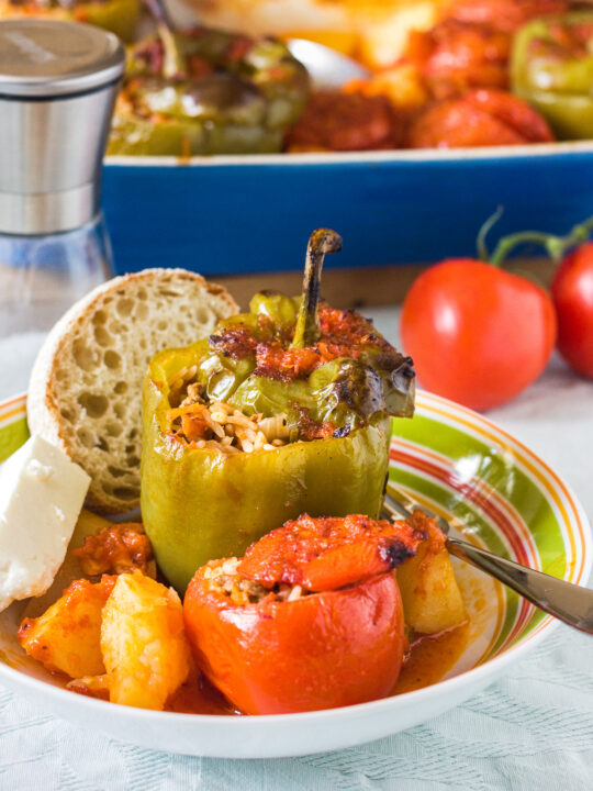 Greek stuffed peppers and tomatoes in a colorful bowl with potatoes, tomato sauce, a chunk of feta cheese and a piece of fresh bread with a blue baking dish of food behind.