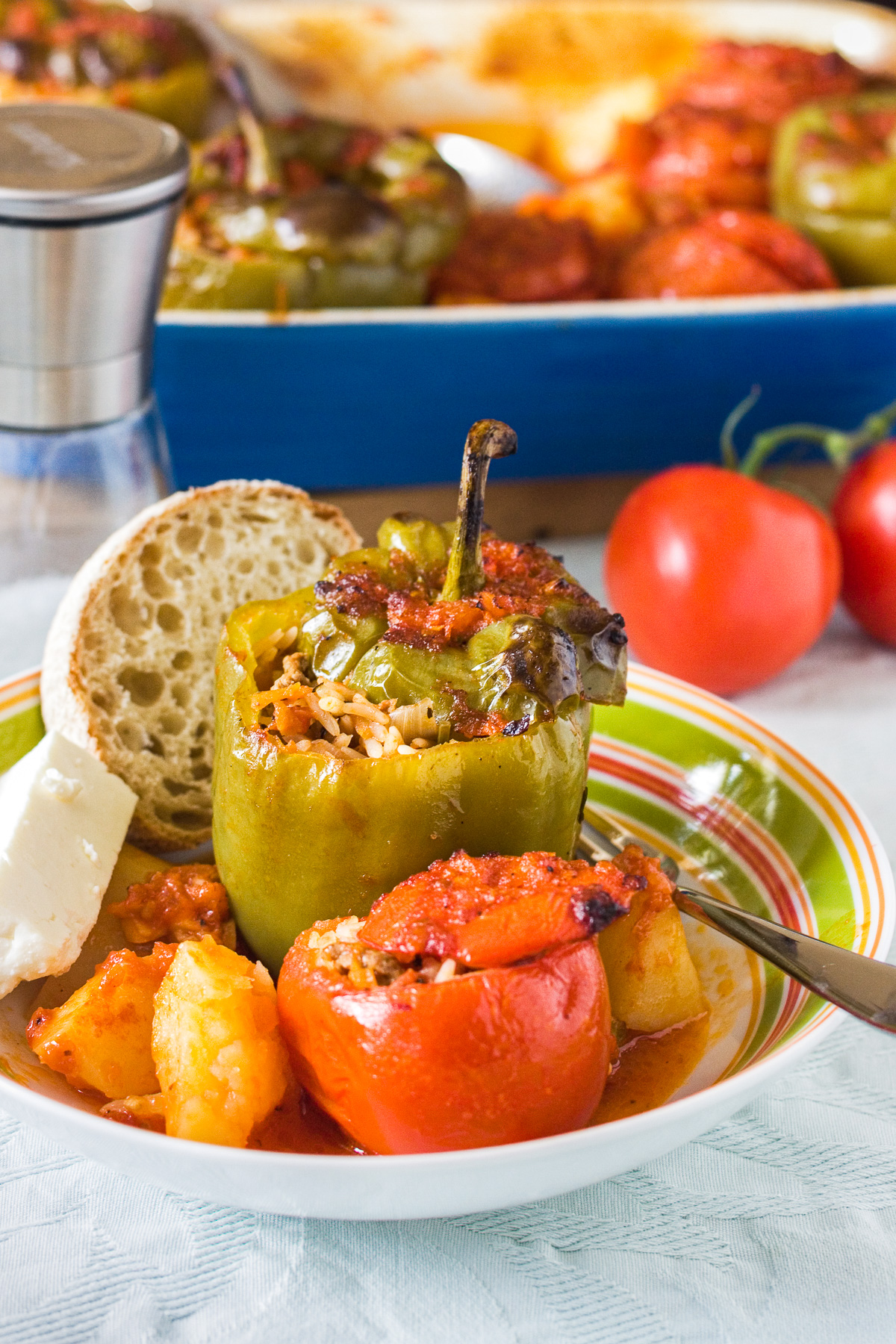 Greek stuffed peppers and tomatoes in a colorful bowl with potatoes, tomato sauce, a chunk of feta cheese and a piece of fresh bread with a blue baking dish of food behind.