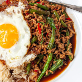 A closeup of a portion of Thai basil pork with rice and an egg on a white plate with a blue rim and with a spoon in it.