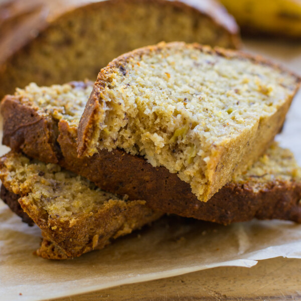 A small stack of slices of air fryer banana bread on a wooden board with the rest of the bread and a banana behind.