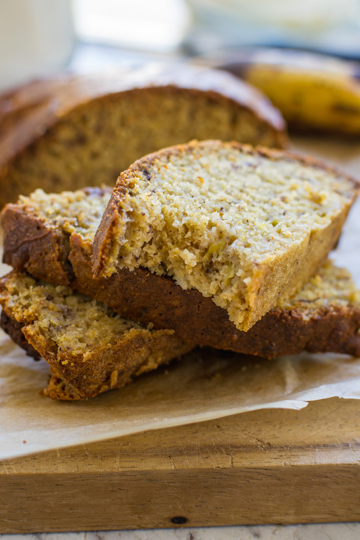 A small stack of slices of air fryer banana bread on a wooden board with the rest of the bread and a banana behind.