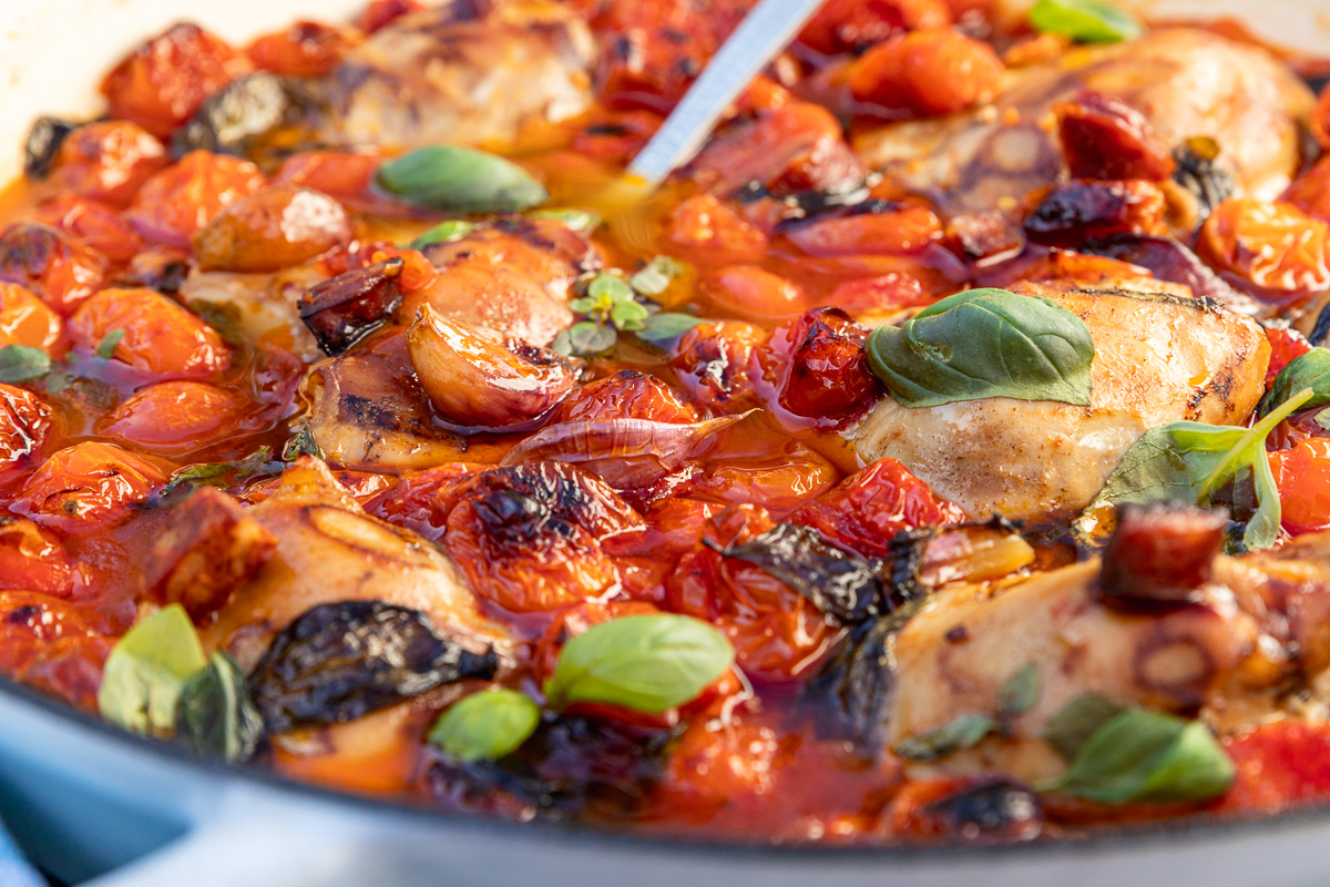 Closeup of baked chicken and chorizo with cherry tomatoes in a blue cast iron pan and with a spoon in it