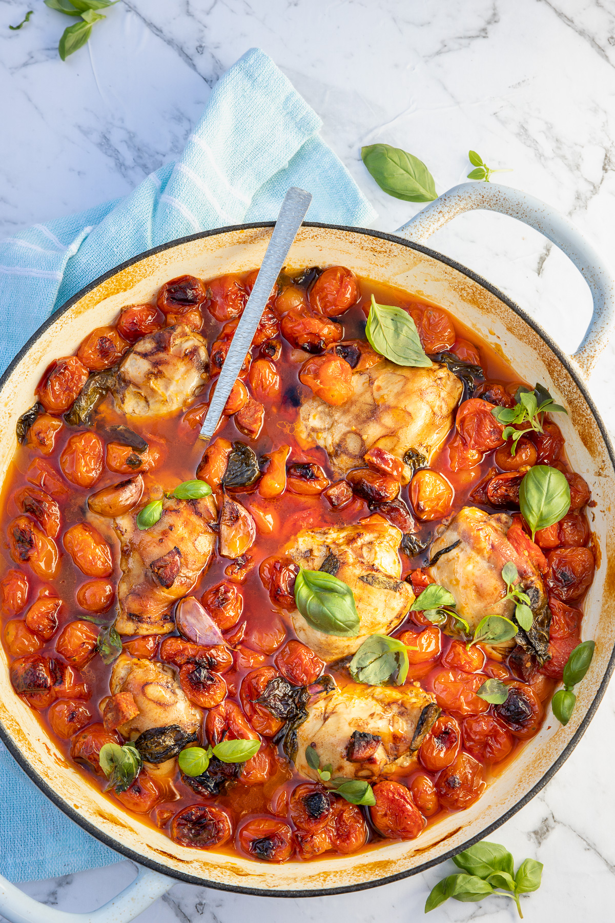 Overhead of a whole blue cast iron pan of chicken and chorizo and cherry tomato bake on a marble background with a blue tea towel.