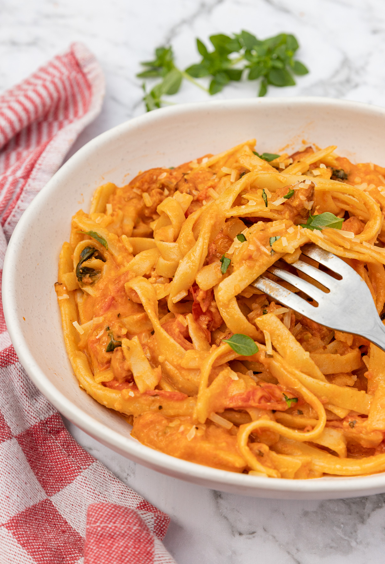 A white dish of creamy chorizo pasta with a fork in it with a red and white checked tea towel and on a marble background.