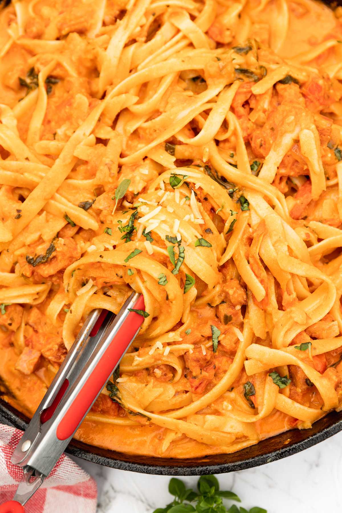 A closeup of a cast iron pan of creamy chorizo pasta with tongs in it and finely chopped basil and parmesan on top.