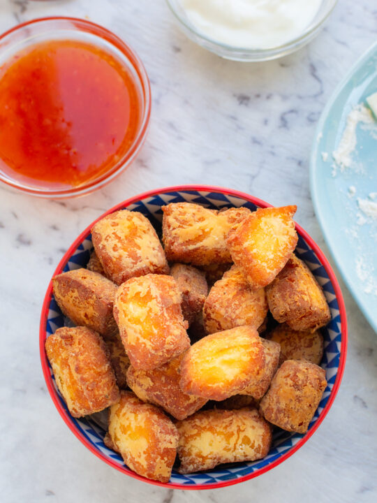 Overhead view of a small blue and red dish of crispy fried halloumi bites with small dishes of dipping sauces in the background.