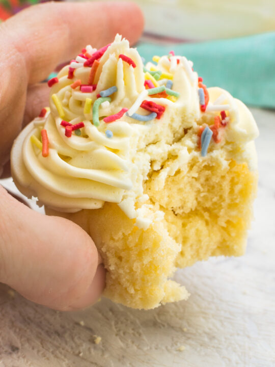 Someone holding up a healthier vanilla cupcake with frosting and sprinkles on it and with a bite taken out of it.