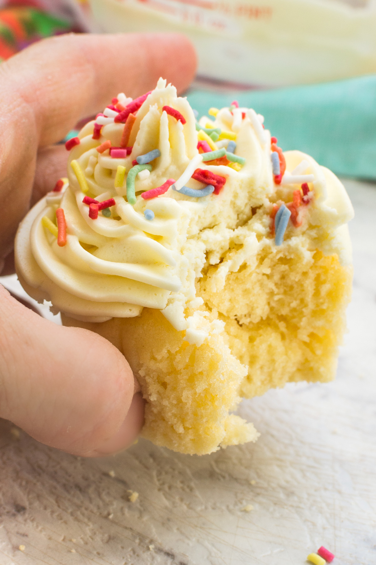 Someone holding up a healthier vanilla cupcake with frosting and sprinkles on it and with a bite taken out of it.