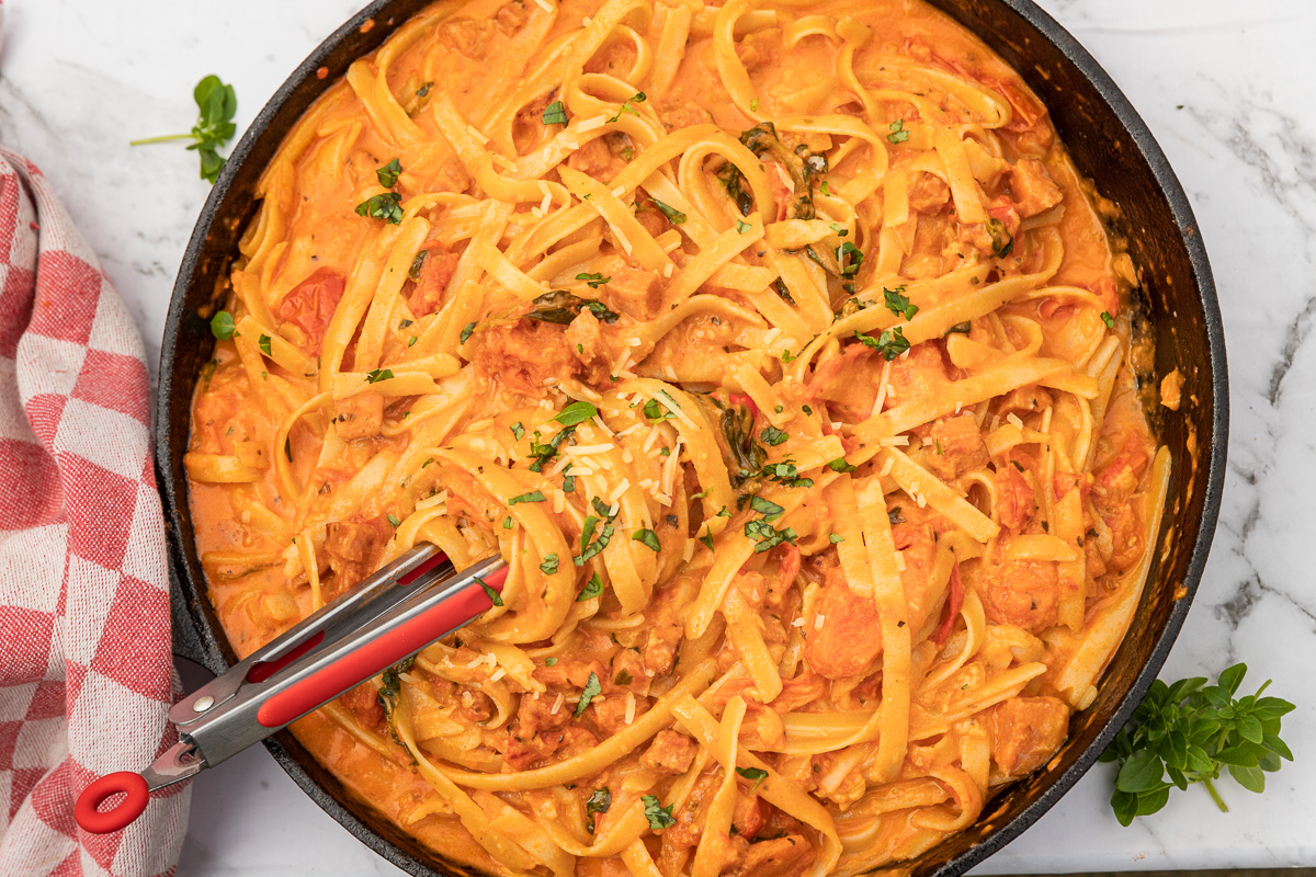 Creamy chorizo pasta in a cast iron pan from above with tongs in it and with a red and white checked tea towel on a marble background.