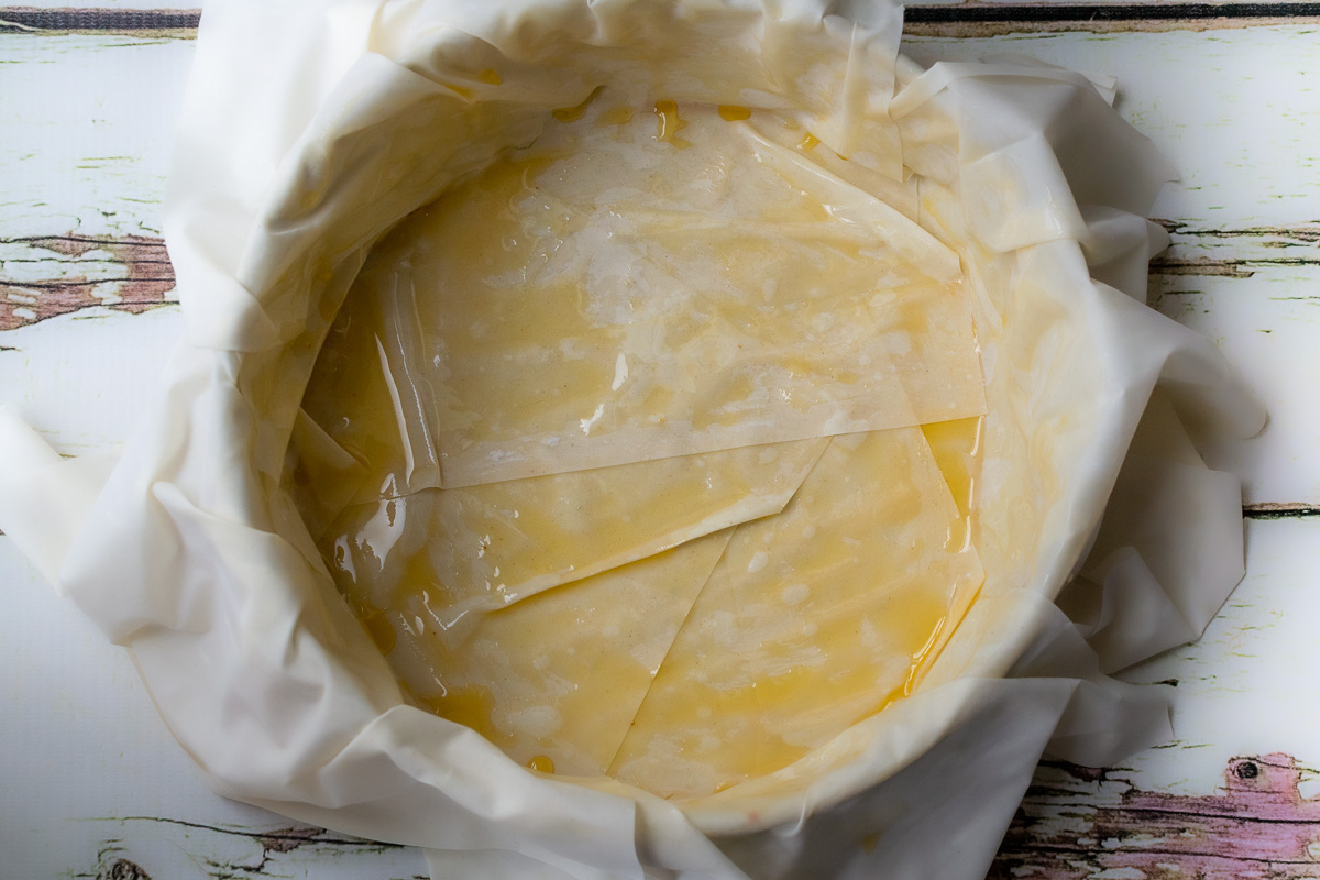 Overhead view of filo pastry layers and olive oil in a springform pan on a white wood background.